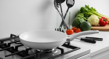 Sartén de cerámica blanca sobre una estufa de gas moderna con quemadores negros. Al fondo, se ven vegetales frescos, incluyendo lechuga, tomates y un pimiento rojo.