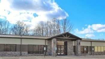 Edificio moderno con fachada de paneles y piedra, entrada acristalada y puertas dobles. Un estacionamiento en primer plano, árboles sin hojas y cielo azul