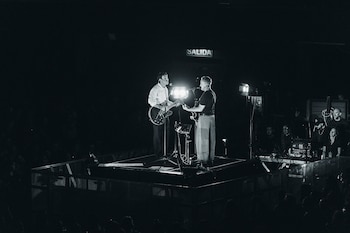 Fotografía en blanco y negro de dos músicos tocando guitarras eléctricas y cantando en un escenario iluminado, con un letrero de 'SALIDA' al fondo y público