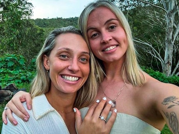 Dos mujeres rubias sonríen a cámara, una con brazo sobre el hombro de la otra y un anillo en la mano. El fondo muestra árboles y vegetación