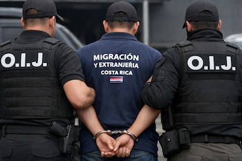 Vista trasera de un hombre esposado con una camisa que dice 'Migración y Extranjería Costa Rica', flanqueado por dos oficiales del OIJ con chalecos negros.