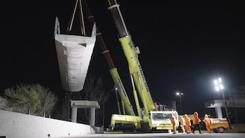 Una grúa amarilla levanta una gran viga de hormigón en la noche mientras varios operarios con uniformes anaranjados observan desde el suelo