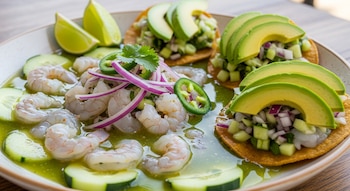 Primer plano de aguachile de camarón con pepino, cebolla morada y cilantro en salsa verde, acompañado de tostadas con aguacate rebanado y limones.