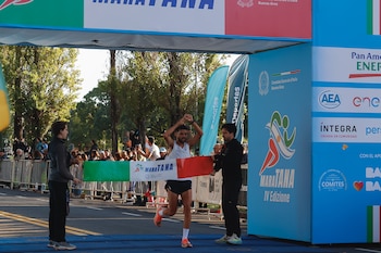 Un corredor masculino con camiseta blanca cruza la meta de la MaraTANA 2026, levantando los brazos. Oficiales y espectadores detrás de las vallas