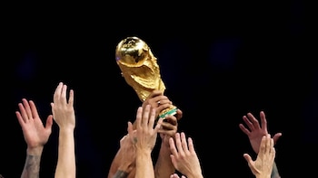Múltiples manos de varias personas levantando el trofeo dorado de la Copa Mundial de la FIFA sobre un fondo oscuro
