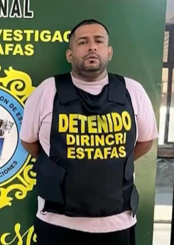Un hombre con barba y camiseta rosa lleva un chaleco negro con la inscripción amarilla 'DETENIDO DIRINCRI ESTAFAS' frente a un fondo verde