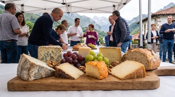 Tabla de madera con varios trozos de queso, uvas rojas y verdes, nueces y pan frente a personas reunidas al aire libre.