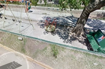 Parque infantil con columpios de cuatro asientos, carrusel, árbol de tronco grueso, dos bancos verdes, sendero de cemento y bordillo