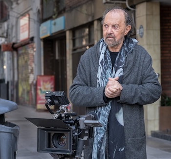 Un hombre con abrigo gris y bufanda de pie en una acera junto a una cámara cinematográfica profesional con parasol y monitor
