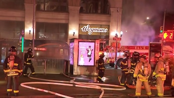 Bomberos uniformados con equipo de protección y mangueras en el suelo frente a un edificio con el letrero Wegmans. Hay humo y camiones de bomberos