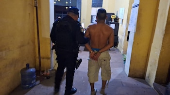 Agente de policía con uniforme oscuro escoltando a un hombre sin camisa con pantalones cortos y esposado por la espalda en un pasillo con paredes amarillas