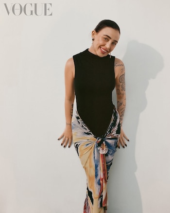 La cantante Nicki Nicole posa sonriendo contra una pared blanca, vistiendo un body negro sin mangas y una falda pareo multicolor atada al frente