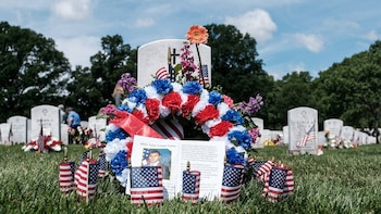 Lápida blanca en cementerio militar adornada con corona de flores rojas, blancas y azules, pequeñas banderas de EE.UU. y foto de soldado en uniforme