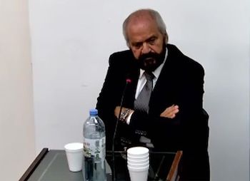 Hombre con barba canosa y traje oscuro sentado en una mesa con micrófono. Una botella de agua y vasos blancos están sobre la mesa