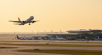 Avión comercial blanco despegando con tren de aterrizaje extendido sobre un aeropuerto moderno con terminal y varias aeronaves en tierra, cielo naranja al atardecer.