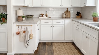 Cocina con gabinetes blancos, encimera de mármol, azulejos tipo metro, utensilios colgados, plantas en maceta y alfombra sobre suelo de madera.
