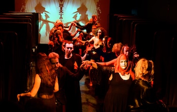 Un grupo diverso de personas, algunas con vestuario teatral, se dan la mano en una formación circular sobre un escenario oscuro con luces rojas y una proyección abstracta de fondo