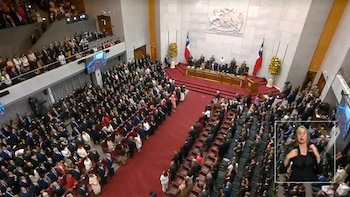 Vista aérea de un gran salón formal con alfombra roja y filas de asistentes. Una tribuna elevada con la bandera de Chile y el escudo nacional al fondo. Intérprete de señas en recuadro