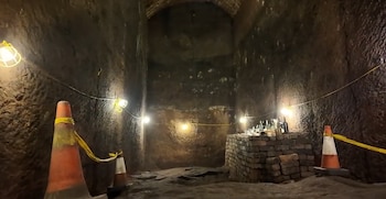 Túnel de piedra con lámparas colgantes, conos de tránsito y una mesa de ladrillos sobre la que se ubican varias botellas de vidrio