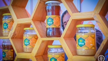 Varios frascos de miel oscura y clara con etiquetas, exhibidos en un estante de madera hexagonal. Un hombre borroso en segundo plano