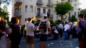 Un grupo de personas en una calle, edificios con balcones al fondo, árboles, vehículos y residuos esparcidos en el suelo