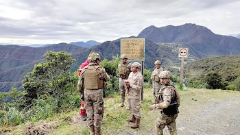 Un grupo de militares con uniformes de camuflaje y cascos de pie en una carretera de tierra, con un cartel y montañas verdes al fondo