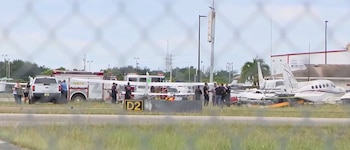 La imagen muestra vehículos de emergencia y aeronaves pequeñas en un aeropuerto. Personal de rescate trabaja en la pista, visible a través de una valla