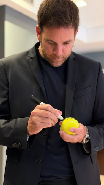 Un hombre vestido con saco oscuro sostiene una pelota de tenis amarilla y escribe sobre ella con un marcador negro