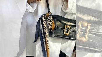 Primer plano de una persona con camisa blanca, bolso negro de mano con textura de cocodrilo y cierre dorado. Un pañuelo de estampado de leopardo y azul está atado a la correa.
