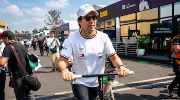 Un hombre con gorra blanca, gafas de sol y camiseta blanca conduce un monopatín en una calle con transeúntes y edificios.