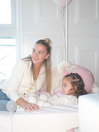 Sofía Calzetti y una niña pequeña están sentadas junto a pelotas blancas y rosadas y globos dentro de un pelotero