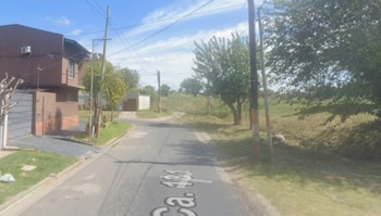 Vista de la calle 131 en Los Hornos, con una casa marrón a la izquierda, postes de luz, árboles y un terreno con pasto y tierra a la derecha