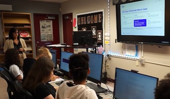 Varios estudiantes trabajan con computadoras en un aula mientras una mujer observa de pie junto a un monitor proyectado