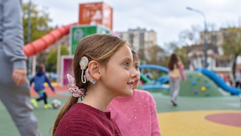 Perfil de una niña con dispositivos auditivos en la oreja y coleta, con toboganes y otros niños jugando al fondo