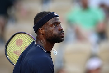 Primer plano de Frances Tiafoe sudando en la cancha, con una diadema azul, camiseta oscura y collar "BIG FOE", y una raqueta de tenis sobre su hombro