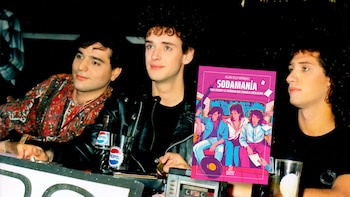 Tres hombres, miembros de Soda Stereo, sentados con fondo oscuro y una mesa con bebidas. Superpuesta está la portada rosa del libro 'Sodamanía'
