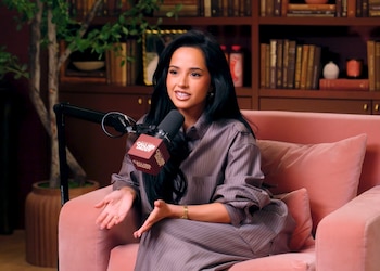 Becky G sentada en un sofá rosa claro, hablando por un micrófono con el logotipo "Call Her Daddy", con una estantería detrás