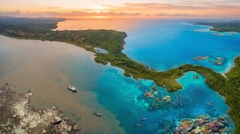 Panorámica aérea de una península densamente arbolada y una bahía con aguas de colores variados. Se observan un barco grande y dos pequeños bajo un cielo anaranjado.