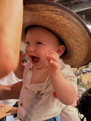 Primer plano de un bebé sonriendo con la boca abierta, lleva puesto un sombrero de paja grande y una camisa clara con tirantes. Brazos de adultos visibles