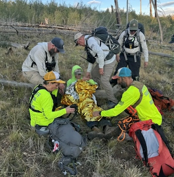 Seis personas, incluyendo a un rescatado envuelto en una manta térmica, rodeadas de mochilas y vegetación en una zona boscosa