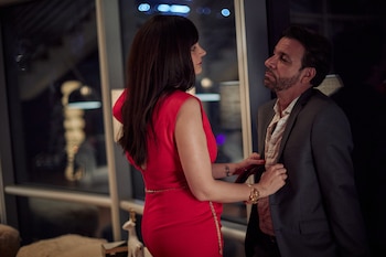 Mujer con vestido rojo y pulsera dorada ajusta la camisa de un hombre con traje oscuro y barba. Se observa un ventanal con luces de ciudad borrosas