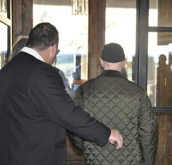 Dos hombres de espaldas. Uno de traje oscuro toca la espalda del otro, quien lleva chaqueta acolchada verde y gorra negra. Puerta y ventanas de madera al fondo
