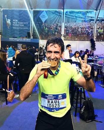 Raúl, un hombre, con camiseta amarilla y dorsal 10525, muerde una medalla de oro. Su mano izquierda hace un signo de victoria. Pancartas de maratón al fondo