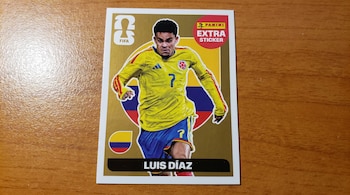 Primer plano de una pegatina Panini "Extra Sticker" dorada con la imagen del futbolista Luis Díaz con el uniforme amarillo y azul de Colombia, sobre una mesa de madera.