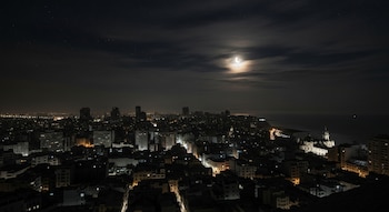 Vista panorámica nocturna de la ciudad de Lima desde arriba, con numerosos edificios iluminados, el océano Pacífico a la derecha y una luna creciente brillante.