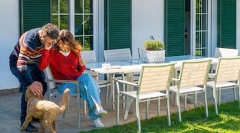 Un hombre acaricia a un perro pequeño cerca de una mujer sentada junto a una mesa blanca en una terraza.
