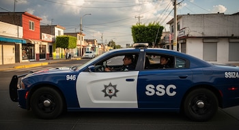 Vista lateral de una patrulla azul y blanca de la SSC con dos policías dentro, circulando por una calle de Tláhuac con casas y negocios a los lados.