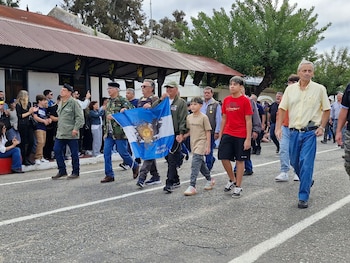 Grupo de veteranos y jóvenes desfilando por una calle con un hombre en el centro llevando la bandera argentina. El cielo está nublado y hay espectadores en los laterales