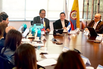 Personas sentadas en una mesa de reuniones con banderas de Colombia y El Salvador, tazas de café, dispositivos electrónicos y cajas azules