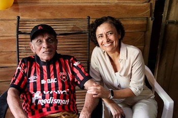 Un hombre mayor con gorra negra y camiseta de fútbol a rayas rojas y negras, y una mujer sonriente de cabello rizado con camisa clara, sentados
